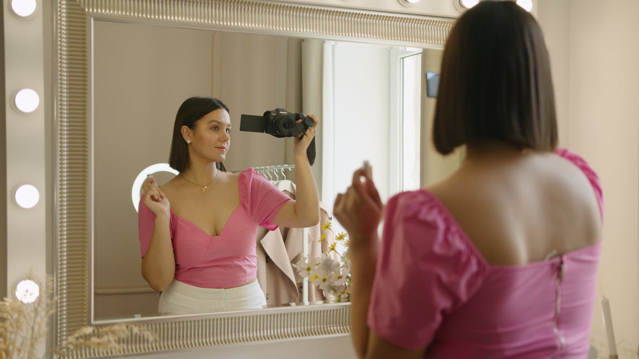 A woman takes a selfie in a well-lit makeup room, wearing a pink top.