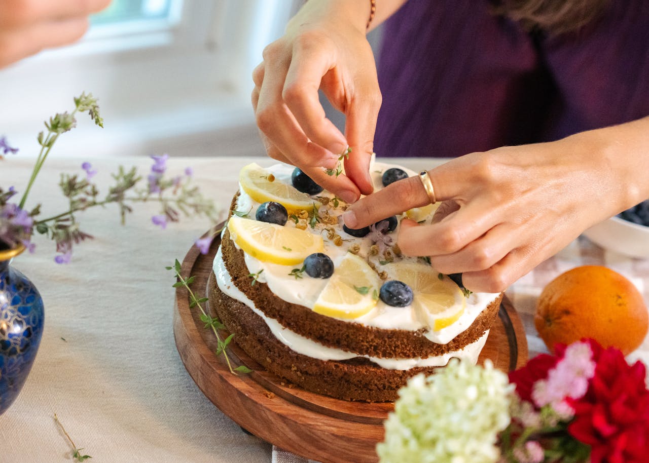 Hands decorating a lemon blueberry cake with herbs, surrounded by fresh flowers.